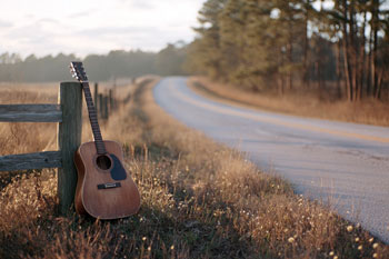 Acoustic guitar leaning against a wooden fencepost beside a country road at golden hour, pine trees in the distance